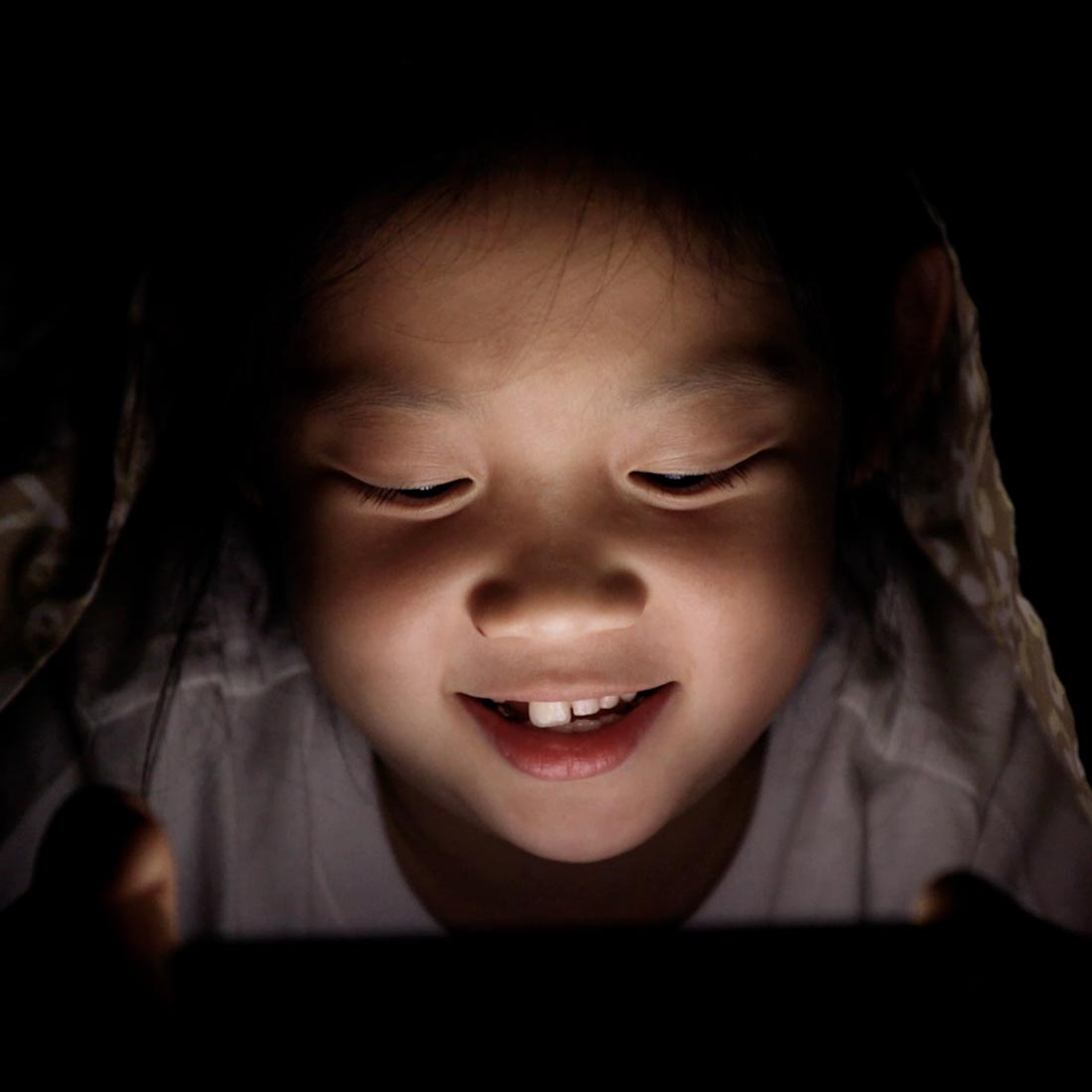 image of a child under of a duvet holding a smartphone and looking at the screen getting his face lighted in the darkness